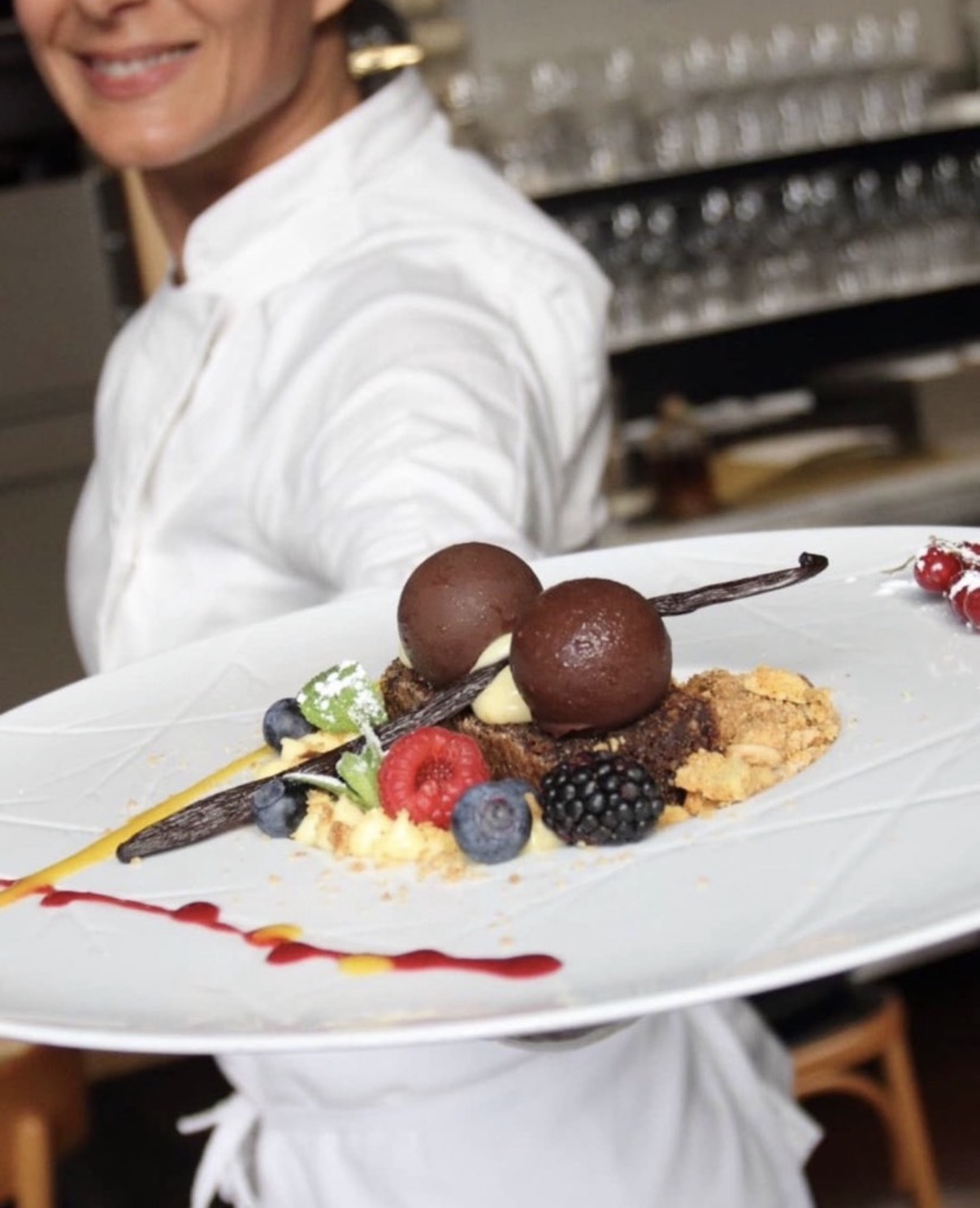 Una persona in divisa bianca porge un piatto con un dessert impiattato e frutti di bosco